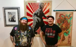 New spaces — Delinquent Gallery & Tattoo KAIJU and Artisan Alley’s Twisted — offer “lowbrow” and boundary-pushing art in Bloomington, says writer Samuel Welsch Sveen. Comics, video-game-themed artwork, cult movies, and tattoos can be found at one; artist studios, a healing shaman, and a retail store for edgier artwork at the other. Pictured here are Delinquent Gallery & Tattoo KAIJU's owners, tattoo artist Chris McVillain, left, and curator Brian Aldridge, right, with art for their inaugural show, "Kai-July." | Photo by Samuel Welsch Sveen