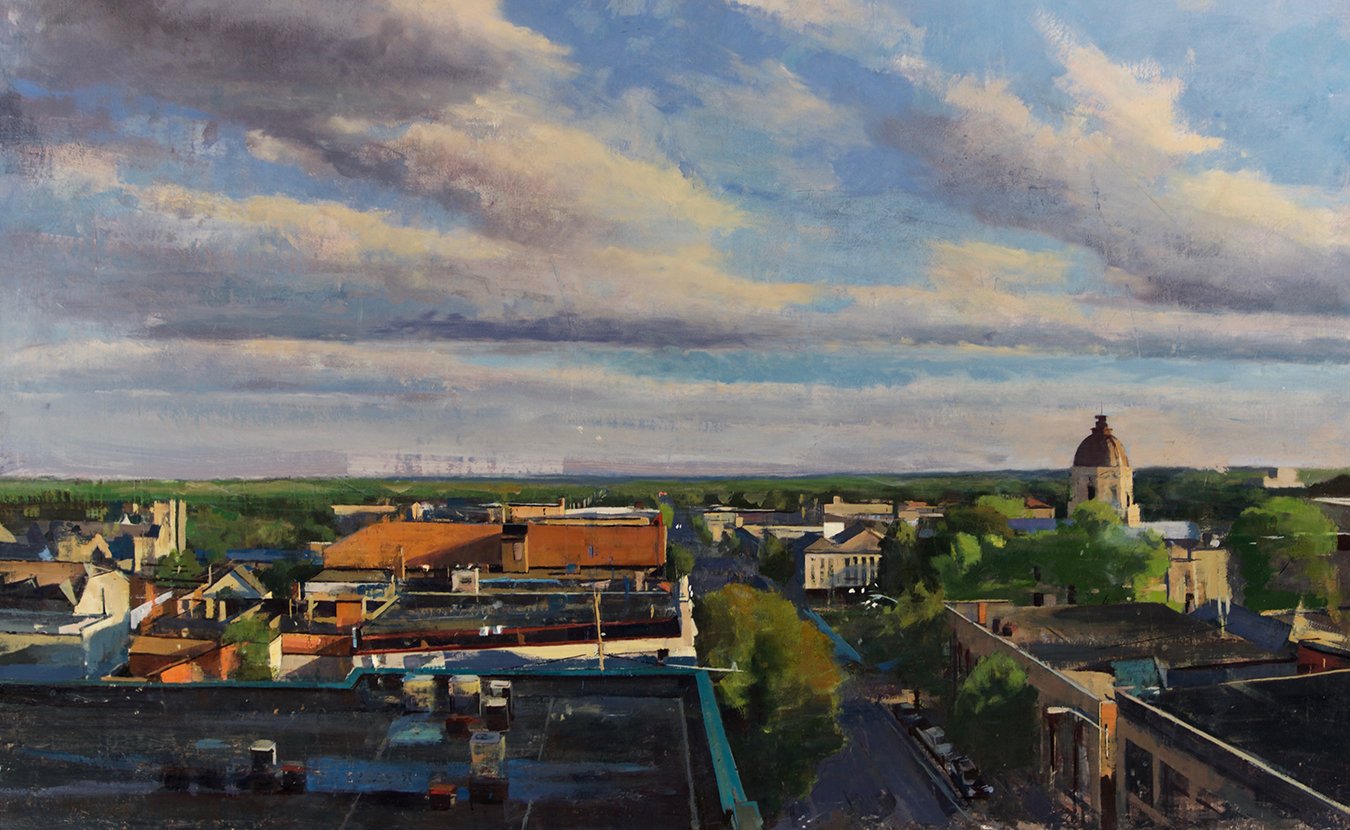 As we enter Thanksgiving holiday week — when “belonging” and feeling “at home” is such a part of the season — writer Amelia Brown describes Bloomington’s “warm feeling of familiarity” and why she belongs here. | ‘Bloomington, View over the rooftops #2,’ Mark W. Ratzlaff, 2016, oil on canvas on board