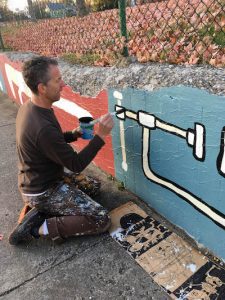 Sam Bartlett renovating his mural at Building Trades Park on West 2nd Street. Painted in 2013, Bartlett’s mural was the first of several murals commissioned by the city. | Photo by M.J. Bower